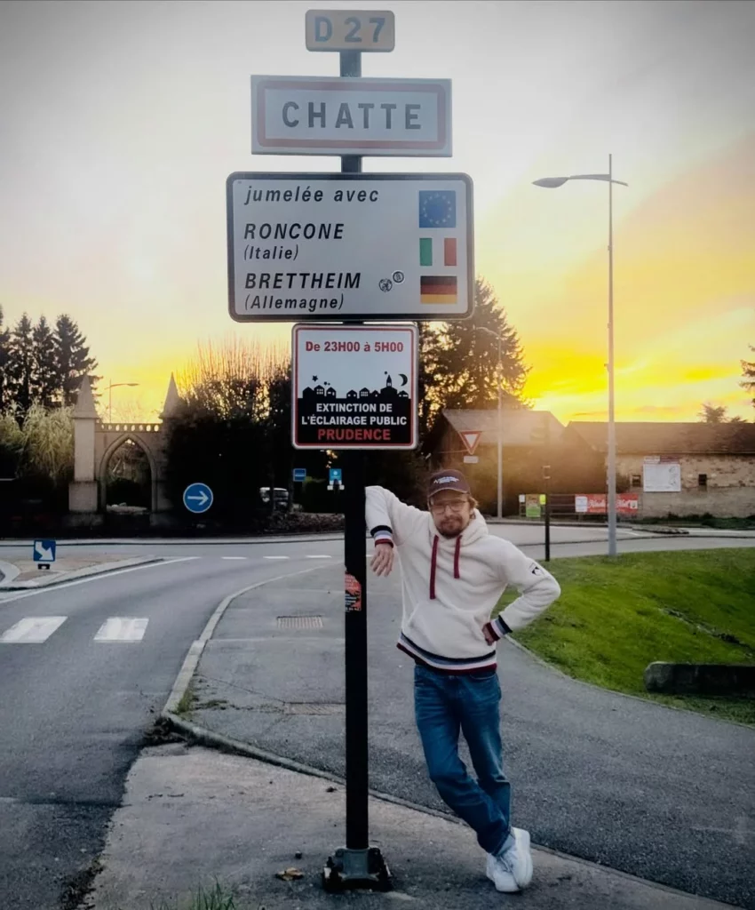 le comédient loràant Deutsch pose devant le panneau de la ville de Chatte en Isère avec un coucher de soleil