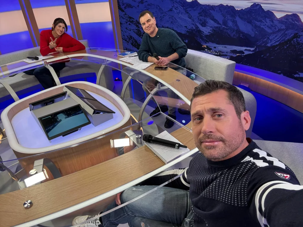 le plateau de France Télévisions avec Guilbaut Colas au premier plan en mode selfie, Laurent Luyat et Ophélie David derrière, plateau des Jeux olympiques de Milano-Cortina