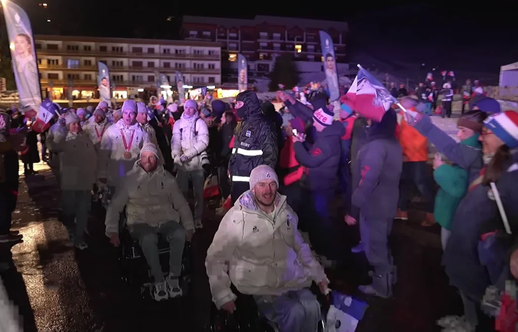 Les athlètes de la délégation française des jeux paralympiques de milano cortina défilent dans la station de charousse la nuit, avec deux athlètes en fauteuil au premier plan